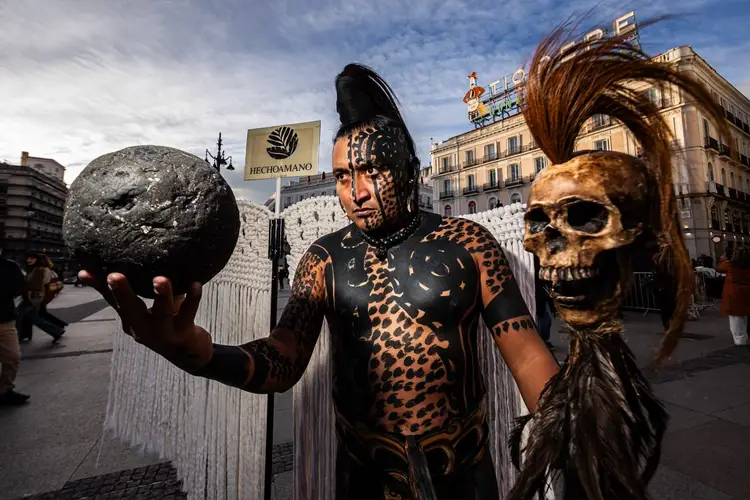 A person in elaborate body paint and costume holds a textured sphere and a skull prop while standing in an urban plaza with classical buildings in the backgroun