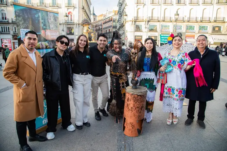 A group of people stand together in an urban plaza in front of classical buildings, with someone wearing traditional embroidered clothing and a pink shawl posin