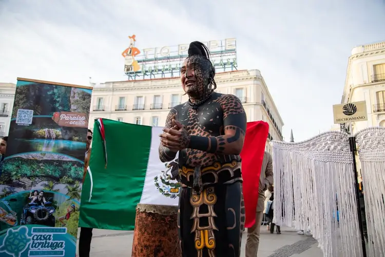 Man with extensive body tattoos holds a Mexican flag while standing in an urban plaza with classical buildings in the background