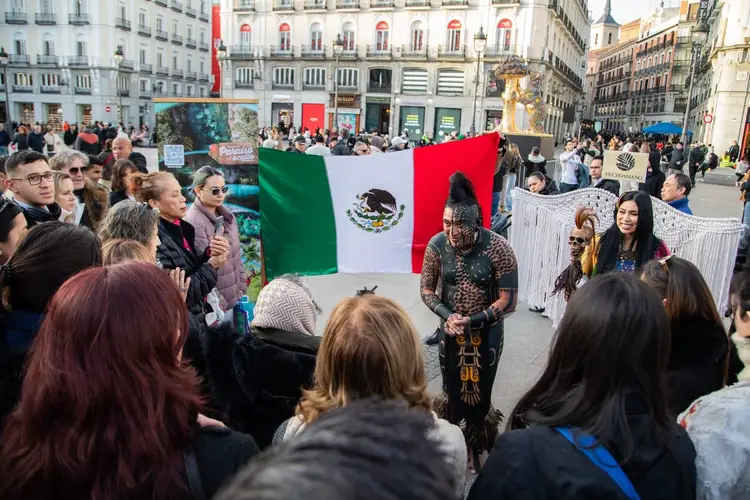 A person in traditional Mayan clothing stands in an urban plaza surrounded by spectators, with a Mexican flag displayed nearby