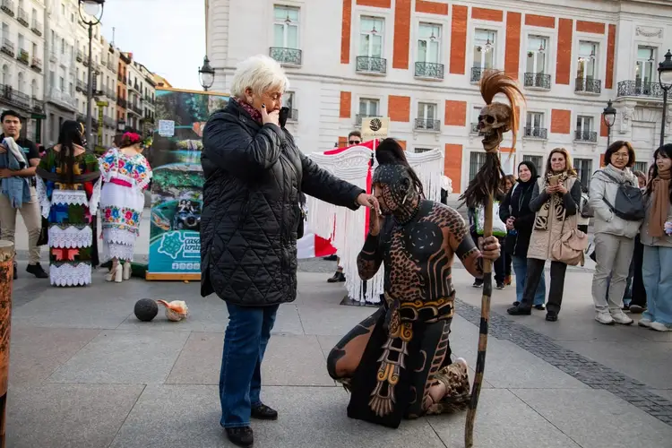 Woman in black jacket interacts with person in elaborate Mayan-inspired costume kneeling in a city plaza surrounded by onlookers