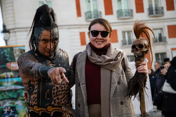 Three people pose together in an urban square, with a person holding a ceremonial staff topped with a skull and feathers standing beside a woman in casual winte