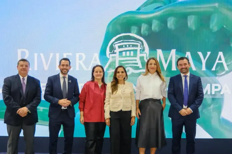 Six people in business attire stand in a line in front of a large screen displaying a turquoise building logo and text Six people in business attire stand in a line in front of a large screen displaying a turquoise building logo and text