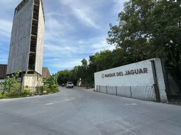 A tall concrete tower and white wall with signage stand along a paved street lined with trees in an urban area