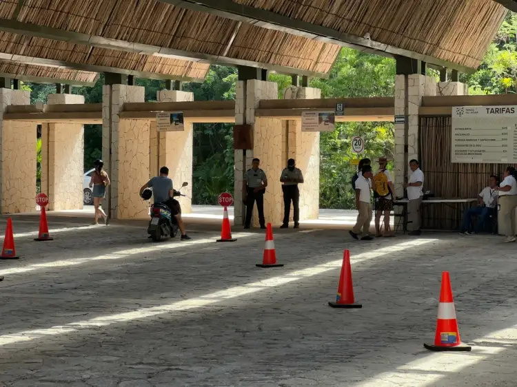 People pass through a covered entrance area with ticket booths and red traffic cones marking a pathway under a thatched roof structure surrounded by greenery