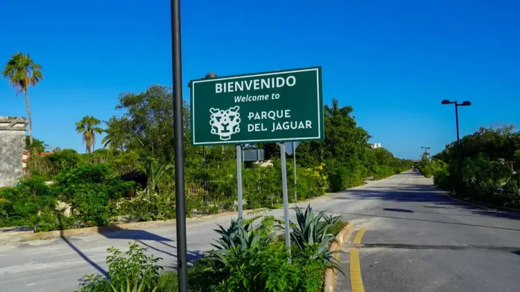 A green welcome sign for a park entrance stands along a paved road lined with tropical vegetation and palm trees under clear blue sky