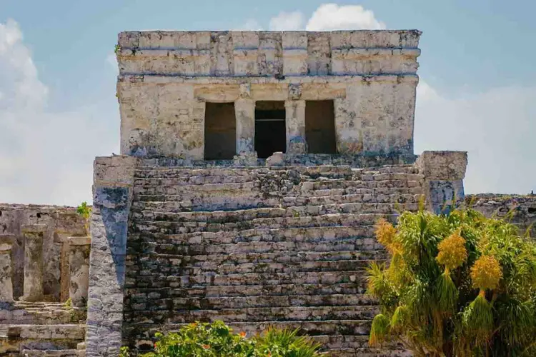 Ancient stone temple structure with three doorways sits atop a stepped pyramid base, with vegetation and cloudy sky visible