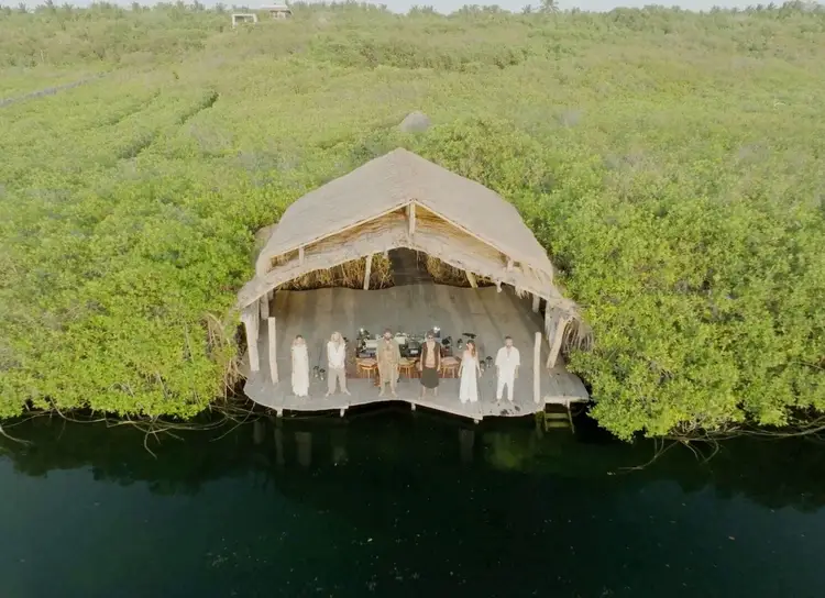 A tent-like structure on the water surrounded by dense green mangrove forest, with silhouettes of people visible inside