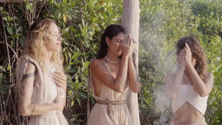 Three women in light-colored draped clothing stand among green foliage and a wooden pole, with two posing thoughtfully and one obscured by haze