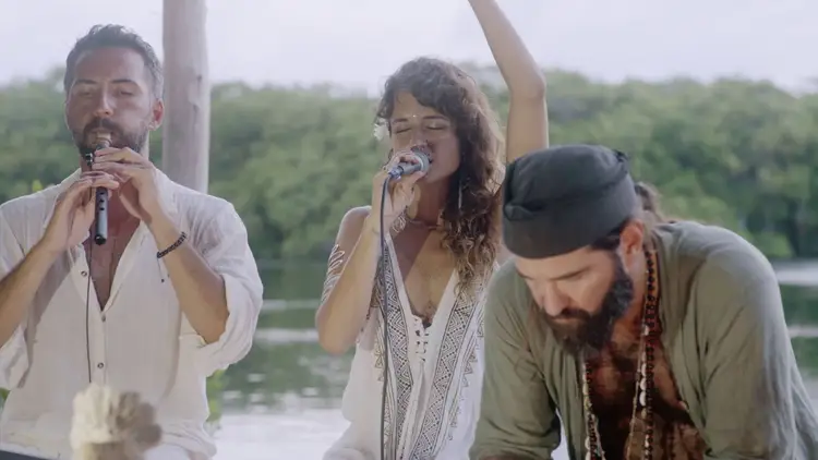 Three musicians perform outdoors with a river and forested shoreline visible behind them, one singing into a microphone while another raises his arm and a third