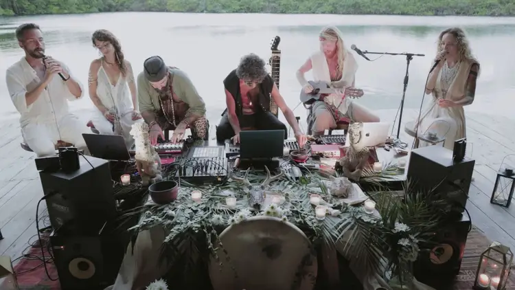 Six musicians perform with laptops and instruments at a table decorated with plants and flowers, positioned on a wooden dock overlooking a calm lake