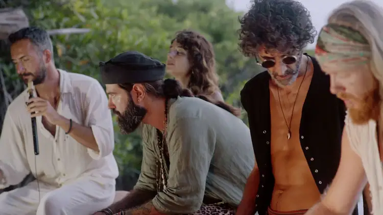 Five musicians sit outdoors with green foliage behind them, one playing a wind instrument while others listen during what appears to be a collaborative performa