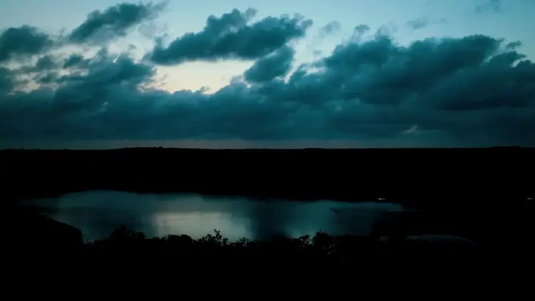 A body of water reflects the evening sky beneath dark clouds, with silhouetted vegetation along the shoreline