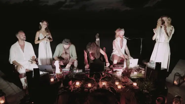 Six musicians perform around a table lit by candles in an outdoor nighttime setting