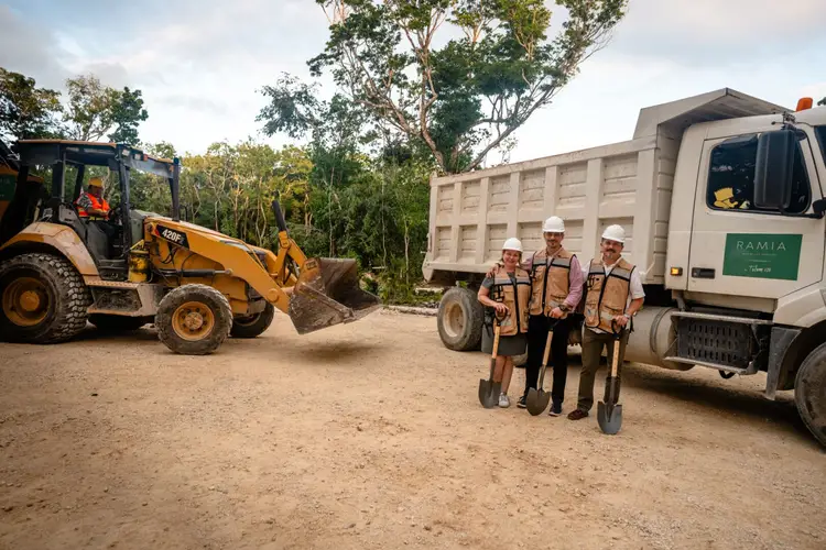 Three construction workers in hard hats and vests stand with shovels in front of heavy machinery and a dump truck at a construction site
