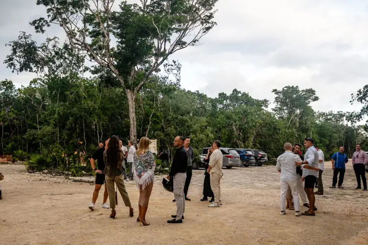 People gather in a cleared dirt area surrounded by tropical forest and parked vehicles