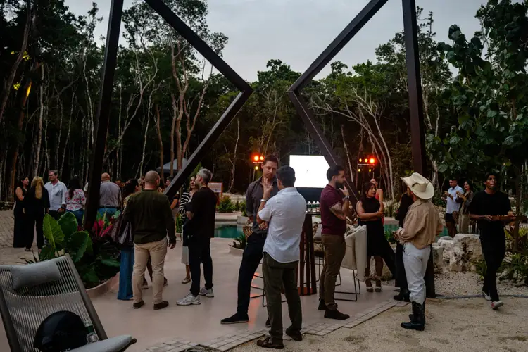 Guests gather at an outdoor event venue with large metal structural beams, surrounded by tall trees and landscaping, as staff and attendees mingle in the evenin
