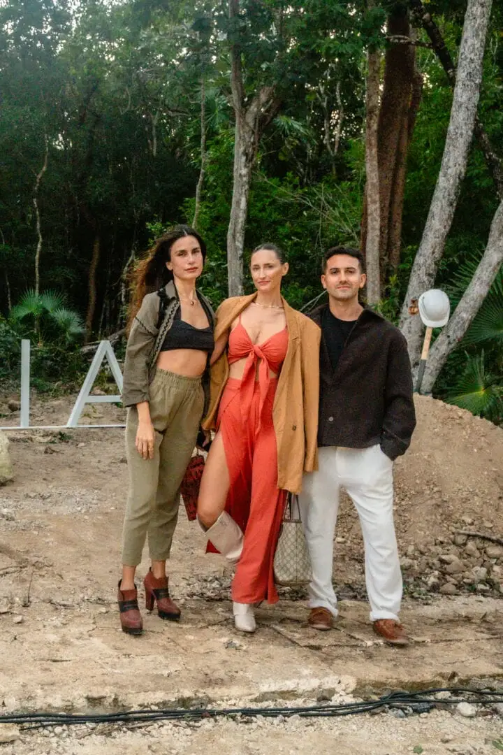 Three people stand on a dirt path among tall trees in a wooded setting