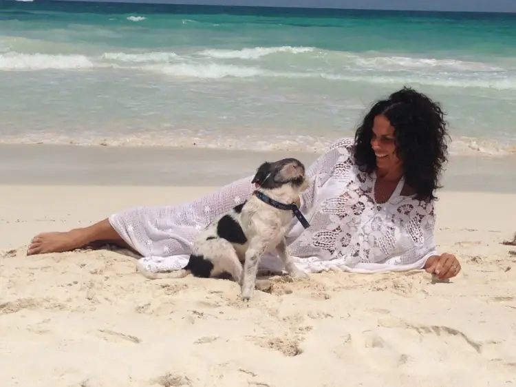 Woman in white clothing lying on sand with a small dog wearing a collar, ocean waves in background