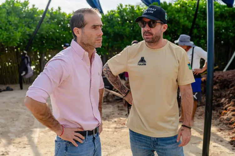 Two men stand on a sandy beach with seaweed and landscaping materials visible behind them, with mangroves and other people in the background