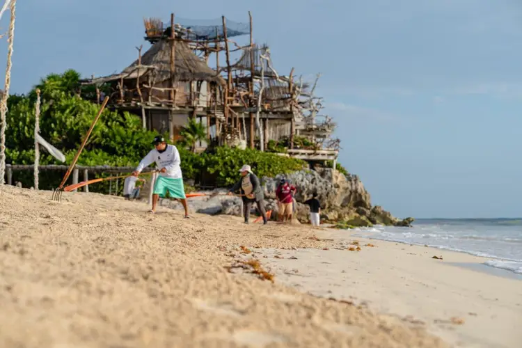 Man playing cricket on a beach with visitors walking nearby and a wooden structure under construction on a cliff in the background