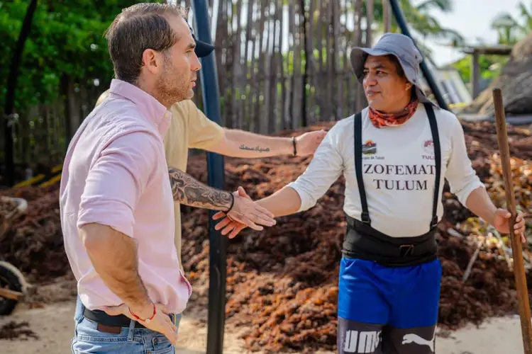 Two men converse while standing in an area with piles of brown seaweed and wooden structures in the background