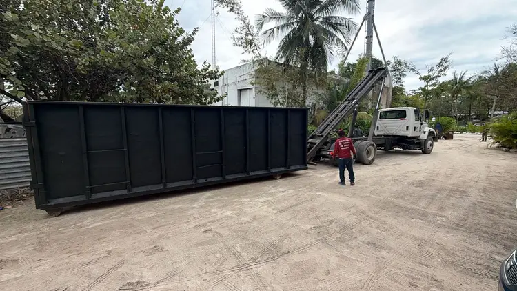 Worker in red shirt stands near a large black waste container being unloaded from a truck in a sandy area with palm trees and buildings in the background