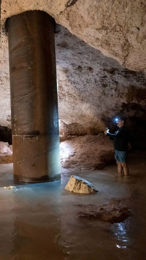 Person with a headlamp examines a large metal pipe in a cave or underground chamber with water on the floor and rock walls Person with a headlamp examines a large metal pipe in a cave or underground chamber with water on the floor and rock walls