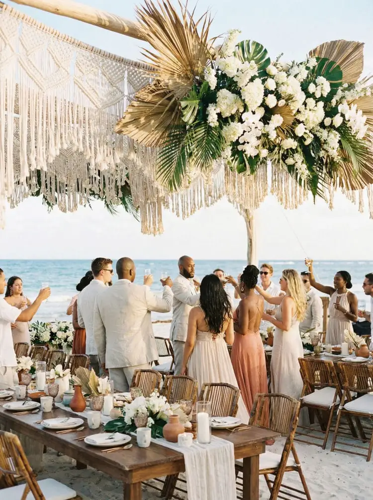 Guests in light-colored clothing gather around a wooden table decorated with white flowers and candles on a beach, standing beneath an ornate hanging floral ins