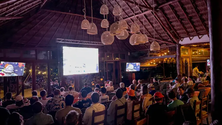 Crowd seated indoors watching a large projection screen under a wooden-beam ceiling with hanging woven pendant lights 1C076135-360A-4664-A275-233F2A70F8A1_1_201_a