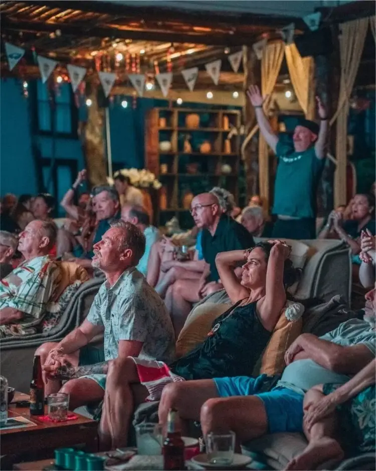 Guests seated on couches watch a game in a decorated outdoor venue with string lights and draped fabric at night 623274829_18552645142004717_5524816242561479821_n