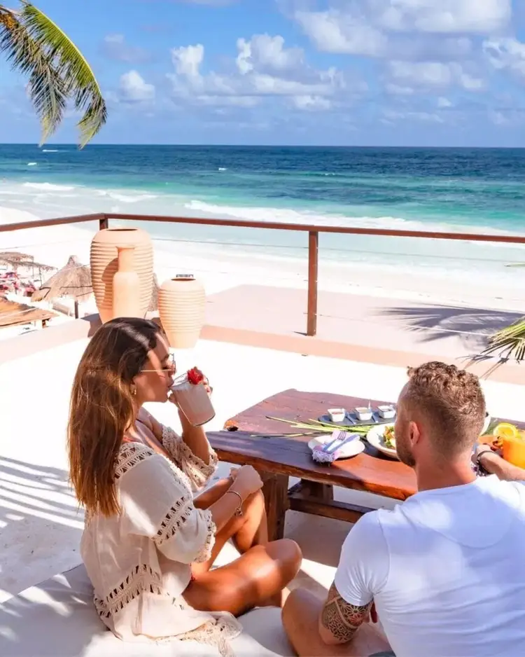 Two people sit at a beachfront table with ocean and palm fronds visible behind them, enjoying food and drinks overlooking the sand and turquoise water 610771721_18549273997004717_3990400888842678869_n