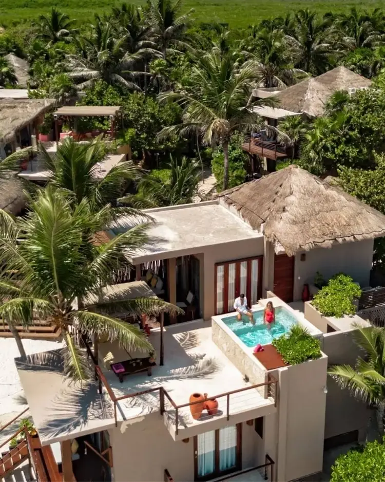 Aerial view of a beachfront resort with thatched-roof bungalows, palm trees, and guests relaxing in a rooftop pool surrounded by tropical vegetation 589355057_18543916813004717_2982633304389818091_n