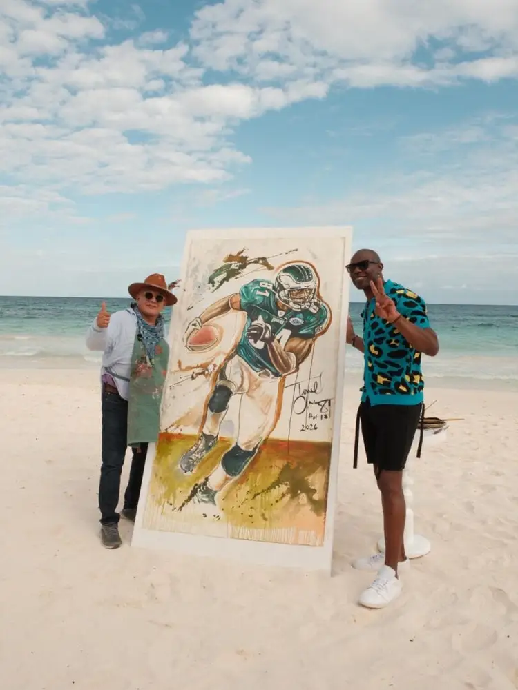Two men flank a large painting of a football player on a beach, with ocean and sky in the background 629575024_18558038134004717_6447698399182641673_n
