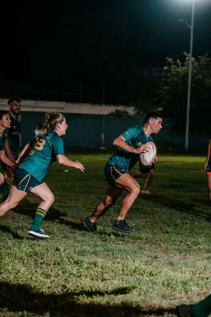 Rugby player carrying the ball runs across a lit field while teammates in matching teal and gold jerseys follow during a night match Rugby player carrying the ball runs across a lit field while teammates in matching teal and gold jerseys follow during a night match