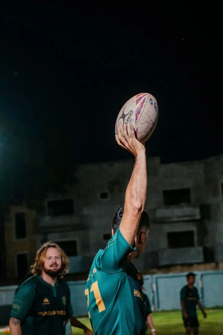 A rugby player in a teal jersey holds a ball high with one arm during a night match, with teammates and a concrete structure visible behind him A rugby player in a teal jersey holds a ball high with one arm during a night match, with teammates and a concrete structure visible behind him