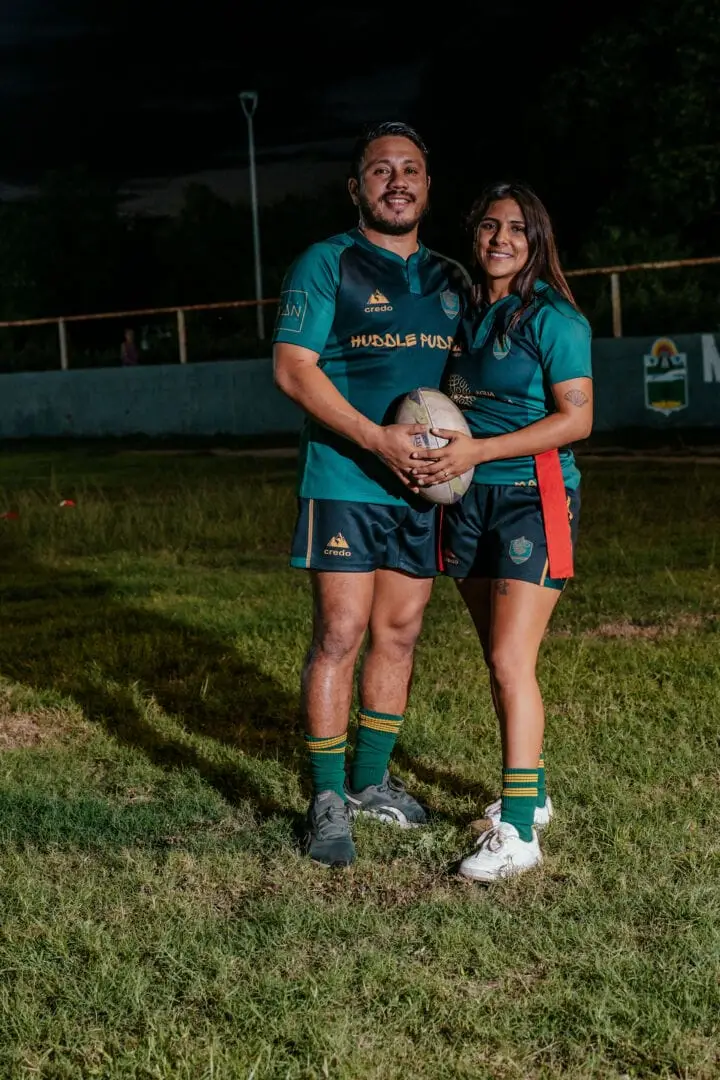 Two rugby players in teal uniforms hold a ball while standing on a grass field at night with stadium fencing behind them Two rugby players in teal uniforms hold a ball while standing on a grass field at night with stadium fencing behind them