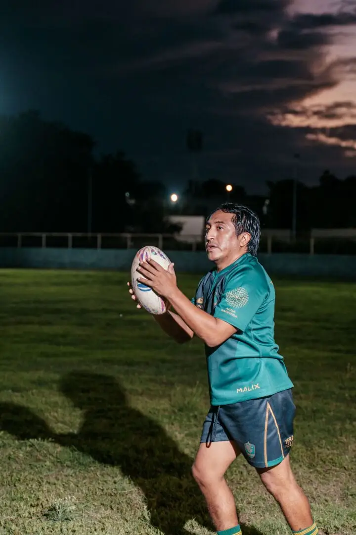 Rugby player in teal jersey holds the ball on a grass field at dusk with stadium lights visible in the background Rugby player in teal jersey holds the ball on a grass field at dusk with stadium lights visible in the background