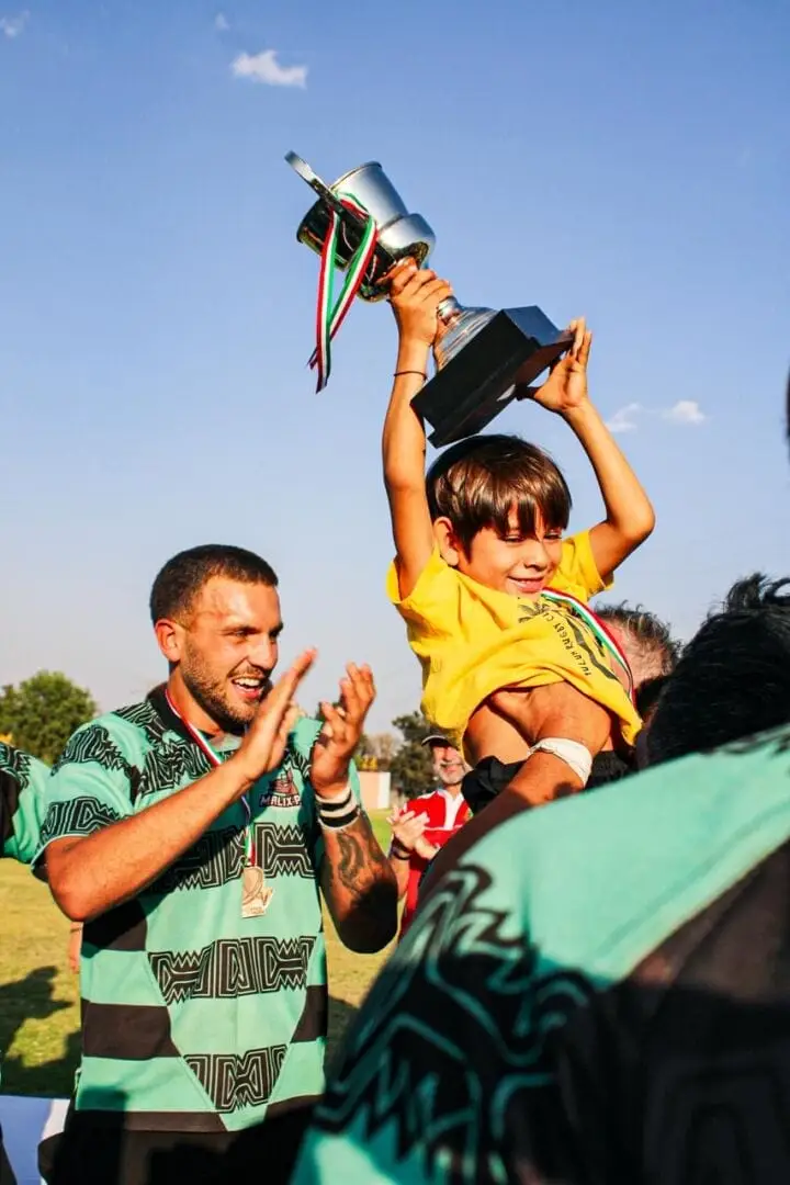 A child in a yellow shirt holds a trophy overhead while a man in a green rugby jersey applauds during an outdoor celebration A child in a yellow shirt holds a trophy overhead while a man in a green rugby jersey applauds during an outdoor celebration