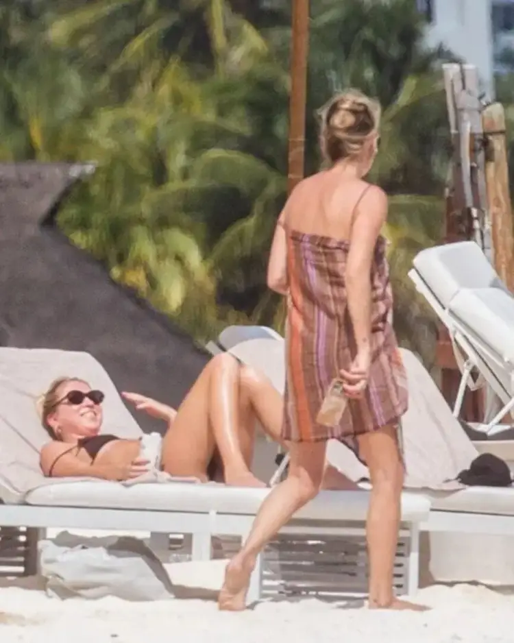 Two people on a beach near white lounge chairs, with one sitting and another standing, palm trees visible in the background HA56u1QWEAAes_v