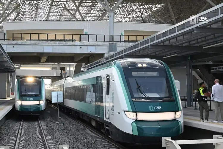 Two teal and white commuter trains are parked at a modern railway station platform with overhead metal lattice structures 560245_123a6c5371d746b182d5e160b8678a7f~mv2