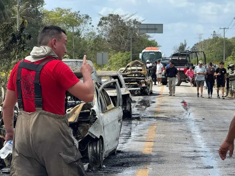 Man in red shirt photographs burned-out vehicle on wet roadway while emergency responders and onlookers gather nearby