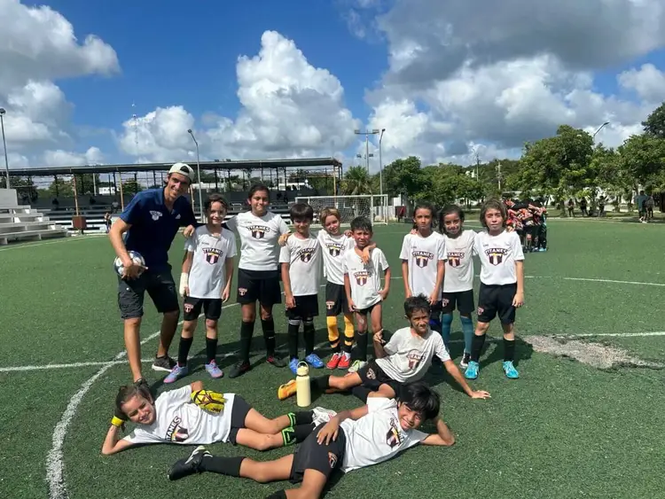 Youth soccer team in matching white and black uniforms poses with their coach on a grass field under partly cloudy skies image13