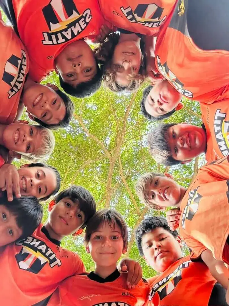 Group of children wearing red soccer team uniforms circle around the camera smiling outdoors beneath a tree image14