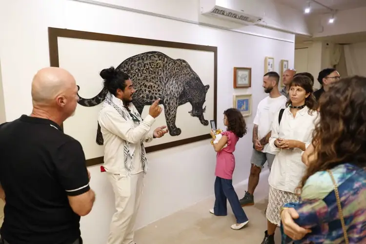 A man in white clothing gestures toward a large framed drawing of a leopard while speaking to a group of visitors in an art gallery _NF_1571