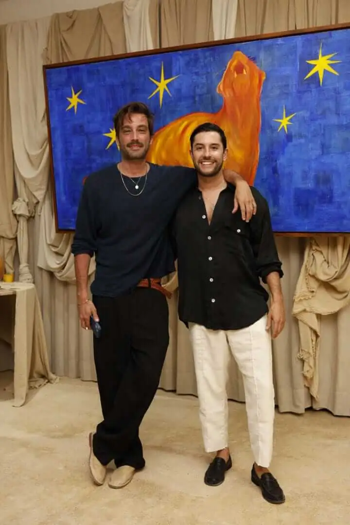 Two men stand together in front of a large painting featuring an orange horse and yellow stars on a blue background, with cream-colored curtains visible behind _NF_1773