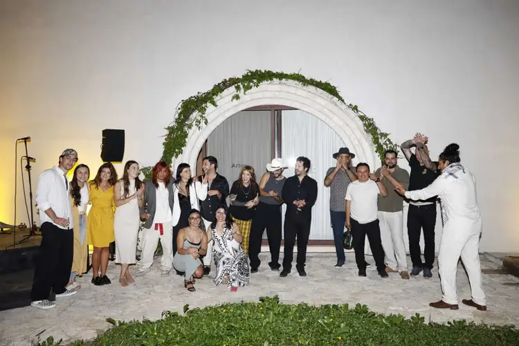 A group of people stand together on sand in front of a white arched structure decorated with greenery, with some wearing formal attire and hats _NF_2467
