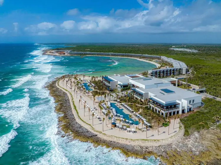Aerial view of a beachfront resort with white buildings, pools, and palm trees on a curved sandy beach with turquoise waves and tropical vegetation beyond 475207018_621469343756430_2029041194972580342_n