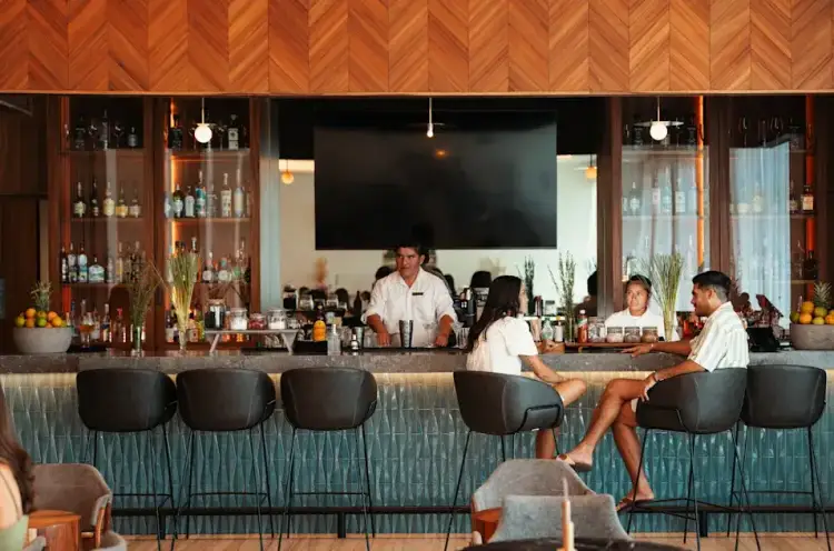 Bartender serves customers at a modern hotel bar with herringbone wood ceiling, black bar stools, and shelves of bottles behind the counter hilton_Muir_day1-1077-Edit