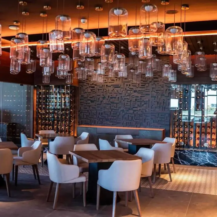 A modern hotel lounge with cream-colored armchairs arranged around wooden tables beneath numerous hanging glass pendant lights with an orange-glowing ceiling 473757732_638065642090507_1882462464234266516_n